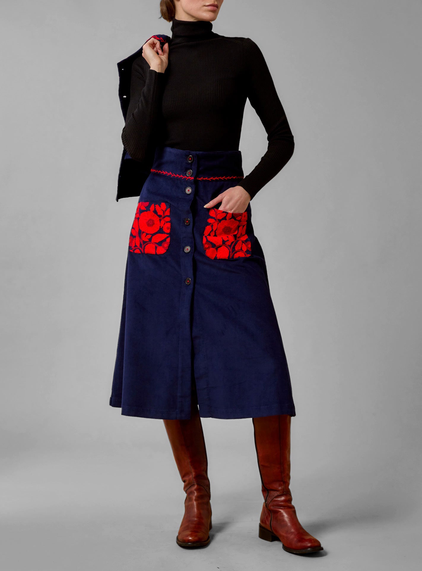 A high-waisted, navy blue cotton corduroy skirt with red wool embroidery and floral pattern on the pockets from Thierry Colson, worn by a model with a turtleneck top.