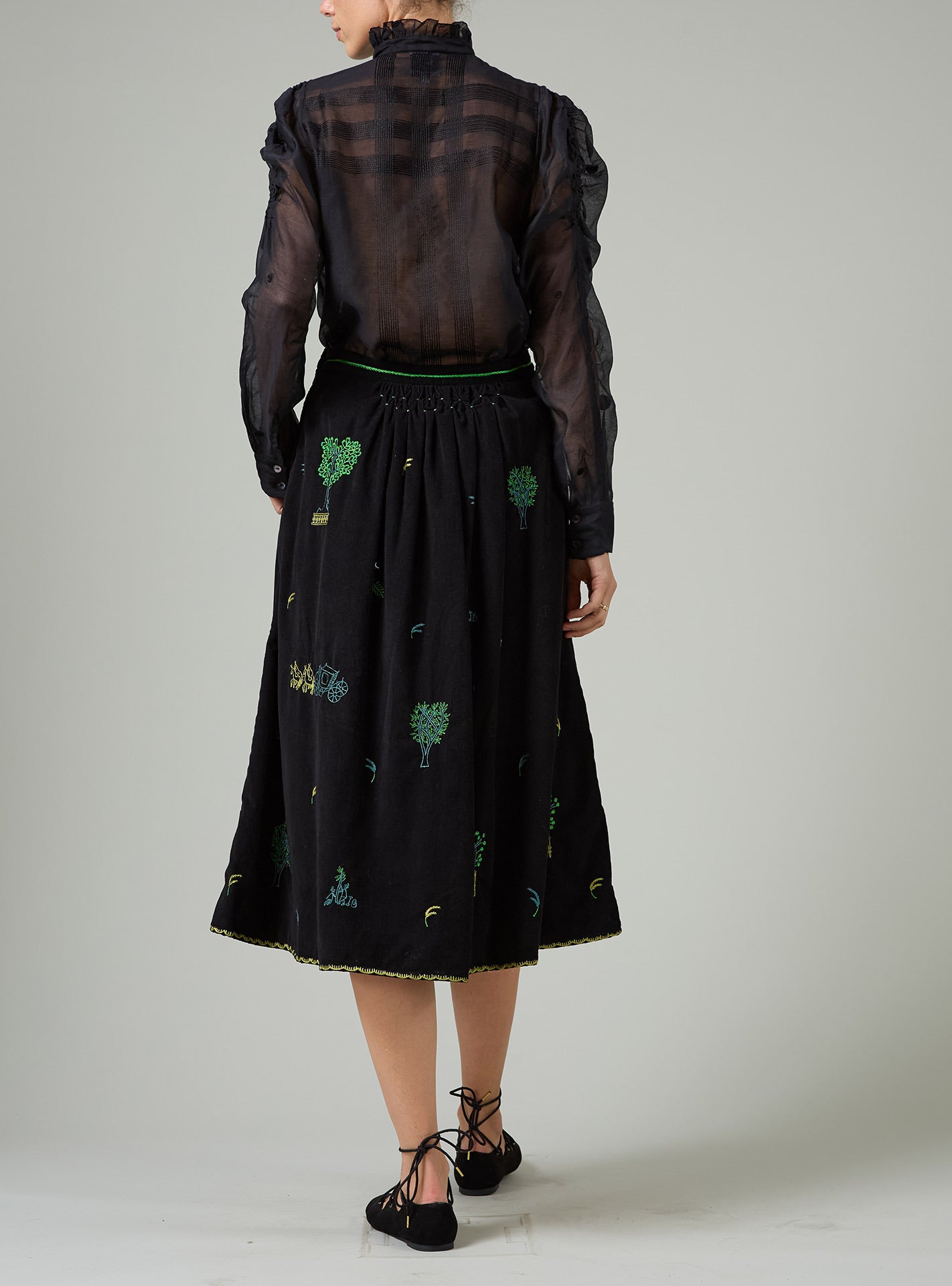 Person wearing a black outfit with a sheer top and embroidered skirt on a plain background