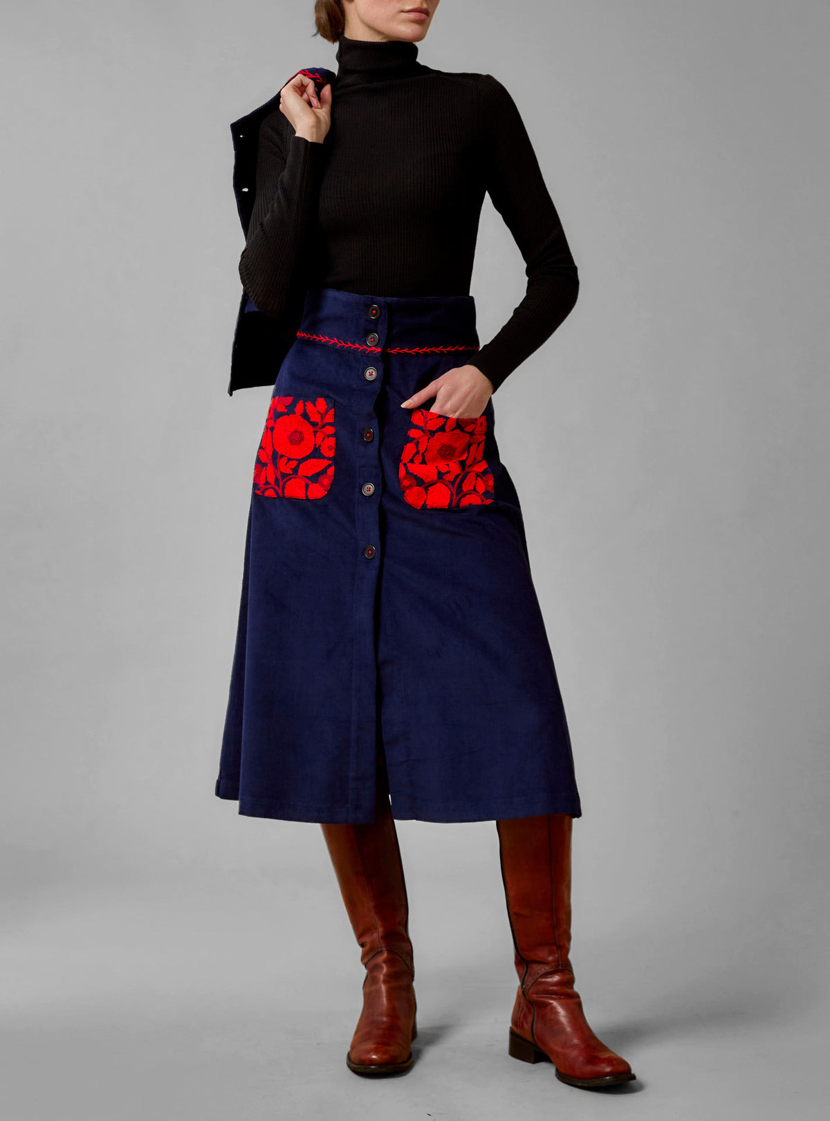 A high-waisted, navy blue cotton corduroy skirt with red wool embroidery and floral pattern on the pockets from Thierry Colson, worn by a model with a turtleneck top.