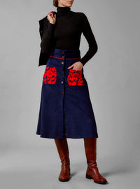 A high-waisted, navy blue cotton corduroy skirt with red wool embroidery and floral pattern on the pockets from Thierry Colson, worn by a model with a turtleneck top.